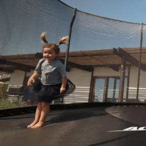Little girl with pigtails in the air jumping on trampoline in a backyard with fence around the trampoline.