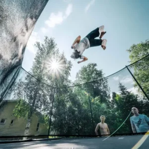 A kid doing a flip on a trampoline with the trees and net behind the child.