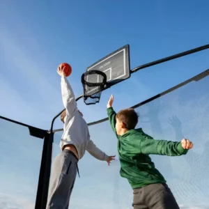 Basketball hoop with net on a trampoline with two kids playing basketball and one almost dunking.