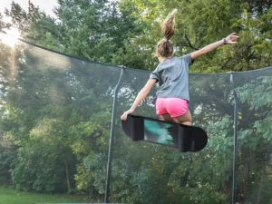 A girl jumping on a trampoline with an acon trampoline skateboard with trees in the background.