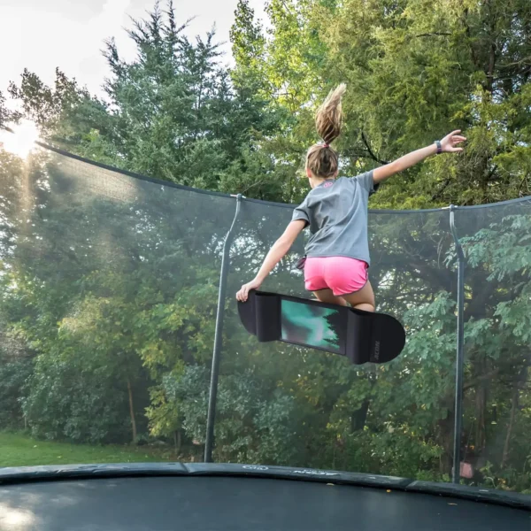 A girl jumping on a trampoline with an acon trampoline skateboard with trees in the background.